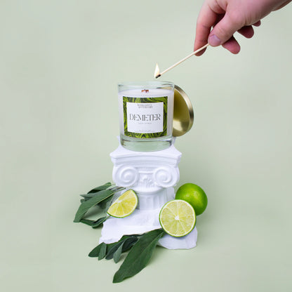 Demeter Candle 8.5 oz being lit with a match on a Greek themed photo set with sliced limes and sage leaves on a soft green background