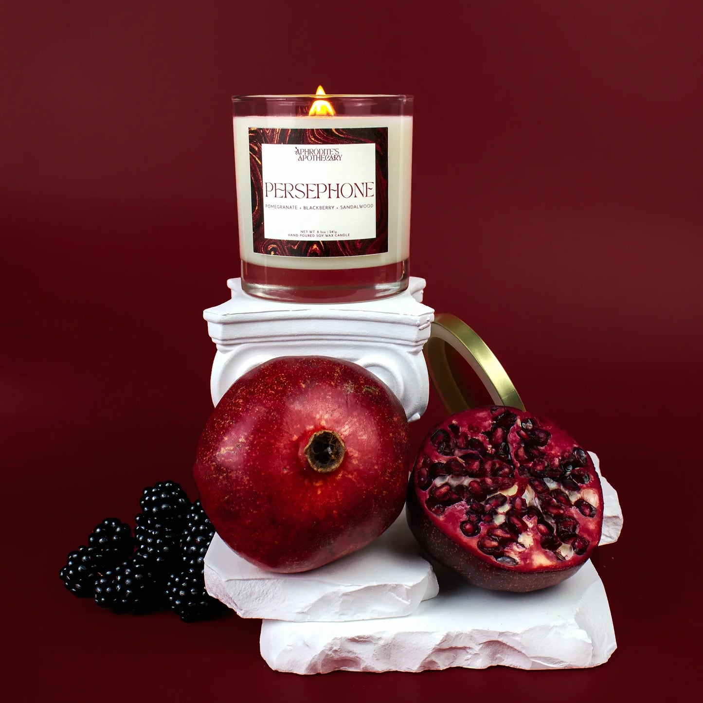 Persephone Candle 8.5 oz on a Greek themed photo set with pomegranates and pomegranate seeds, and blackberries on a deep red background.